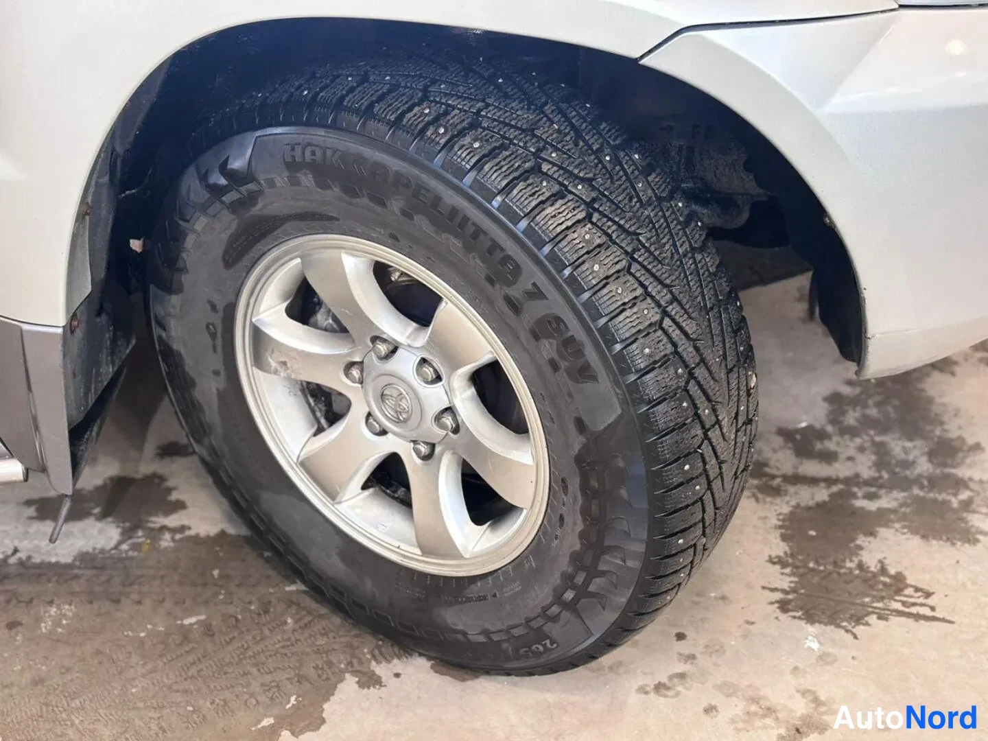Muu: Lähikuva ajoneuvon pyörästä: hopeavanteinen Toyota-merkitty vanne ja naulattu Nokian Hakkapeliitta 7 -talvirengas, märällä lattialla.