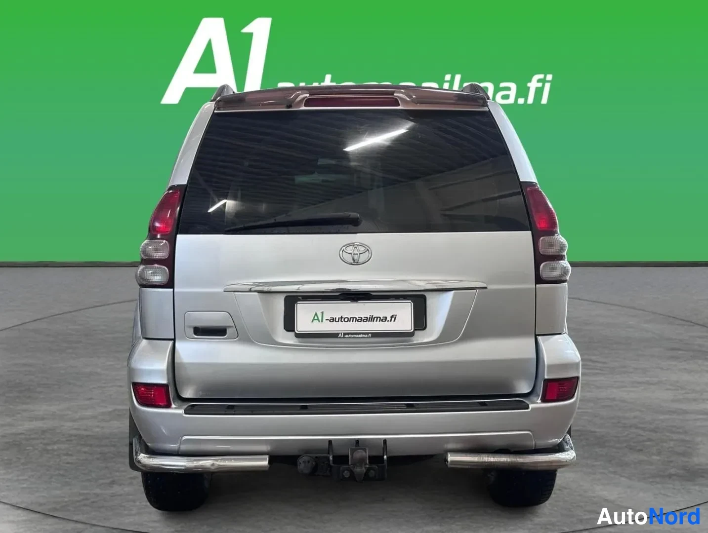 Takaa: Hopeanvärinen Toyota Land Cruiser suoraan takaa kuvattuna; näkyvät takavalot, Toyotan merkki, rekisterikilven paikka A1-automaa ilma.fi -kehyksessä sekä takana