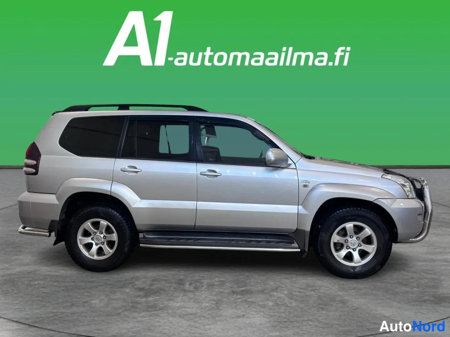 Sivulta: Hopeanvärinen Toyota Land Cruiser 120 kyljestä sivulta kuvattuna; näkyvät ovet, mustat ikkunat, kattotangot, sivulaitetangot ja konepellin etunenän ilmasuodatin