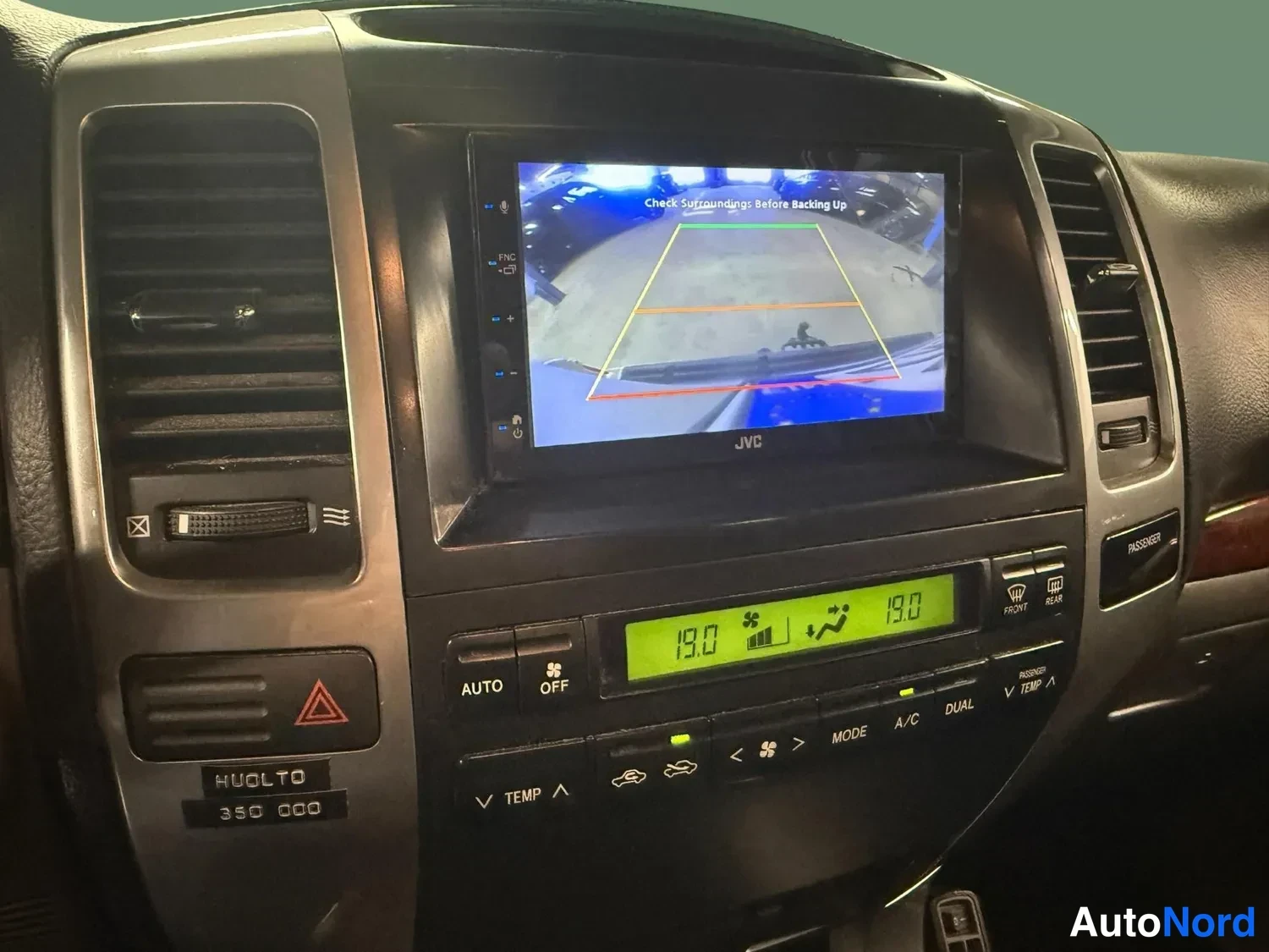 Muu: Kojelaudan keskiosa: JVC-näyttö näyttää peruutuskameran kuvan tekstillä 'Check Surroundings: Before Backing Up', alla ilmastoinnin hallinta näytöllä 19.0, AUTO-