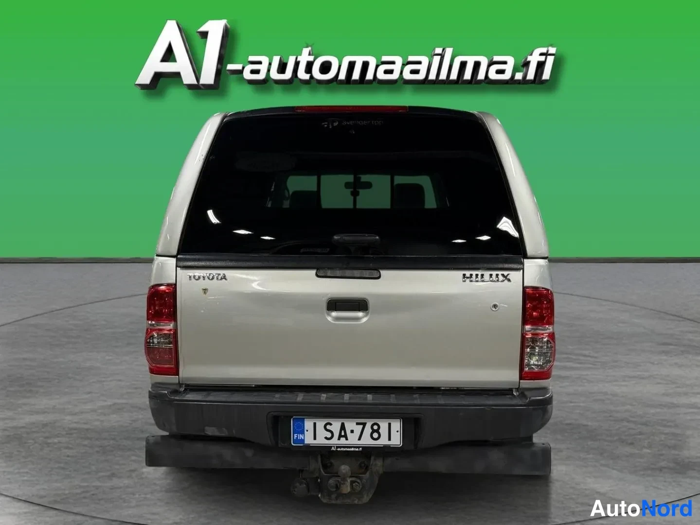 Takaa: Toyota Hilux -nova takaa kuvattuna: hopeanbeige koriratkaisu ja selkäkansi, Toyota- ja Hilux-tekstit sälekaihtimessa, rekisterikilpi ja vetokoukku.