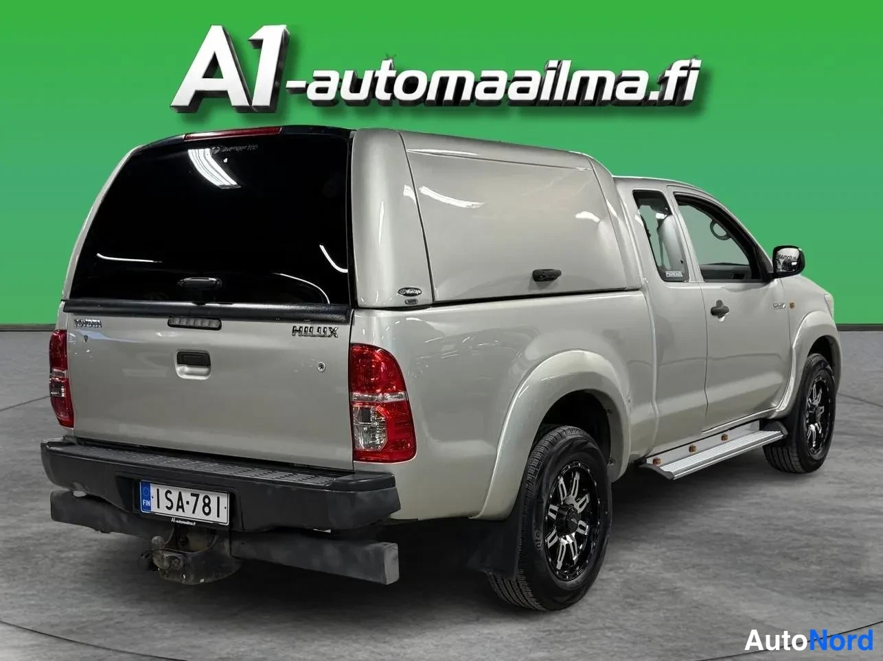Takaviisto: Toyota Hilux -nova takaosan ja oikean sivun suunnasta kuvattuna: hopeanbeige sälekansi korilla, tummat kevytmetallivanteet ja oven alapuolinen askelma näkyvät.