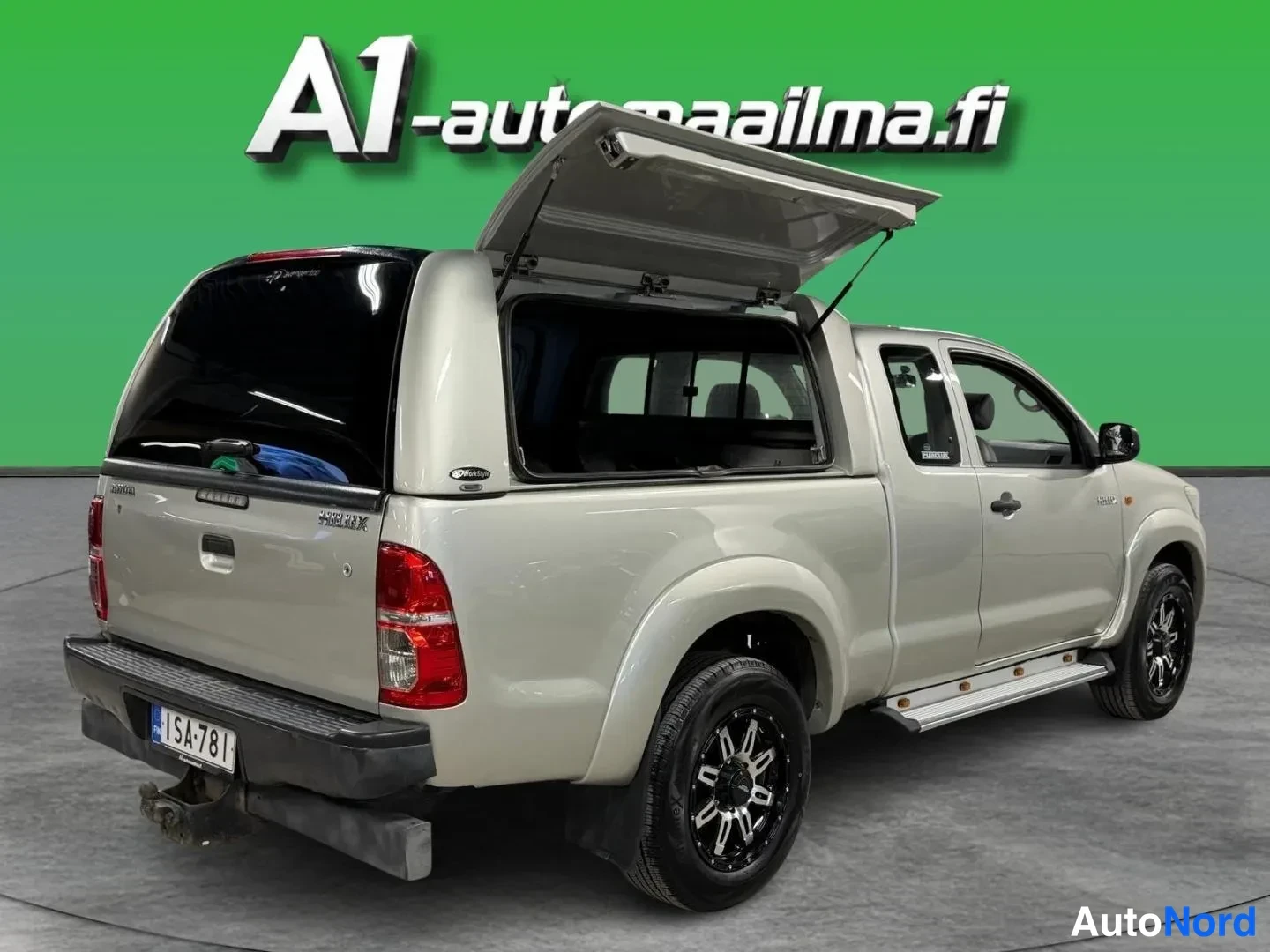 Takaviisto: Toyota Hilux -nova takaosan ja oikean sivun suunnasta kuvattuna: korin takaluukki on auki ylöspäin ja lavan sivuikkuna näkyy, hopeanbeige koriratkaisu.