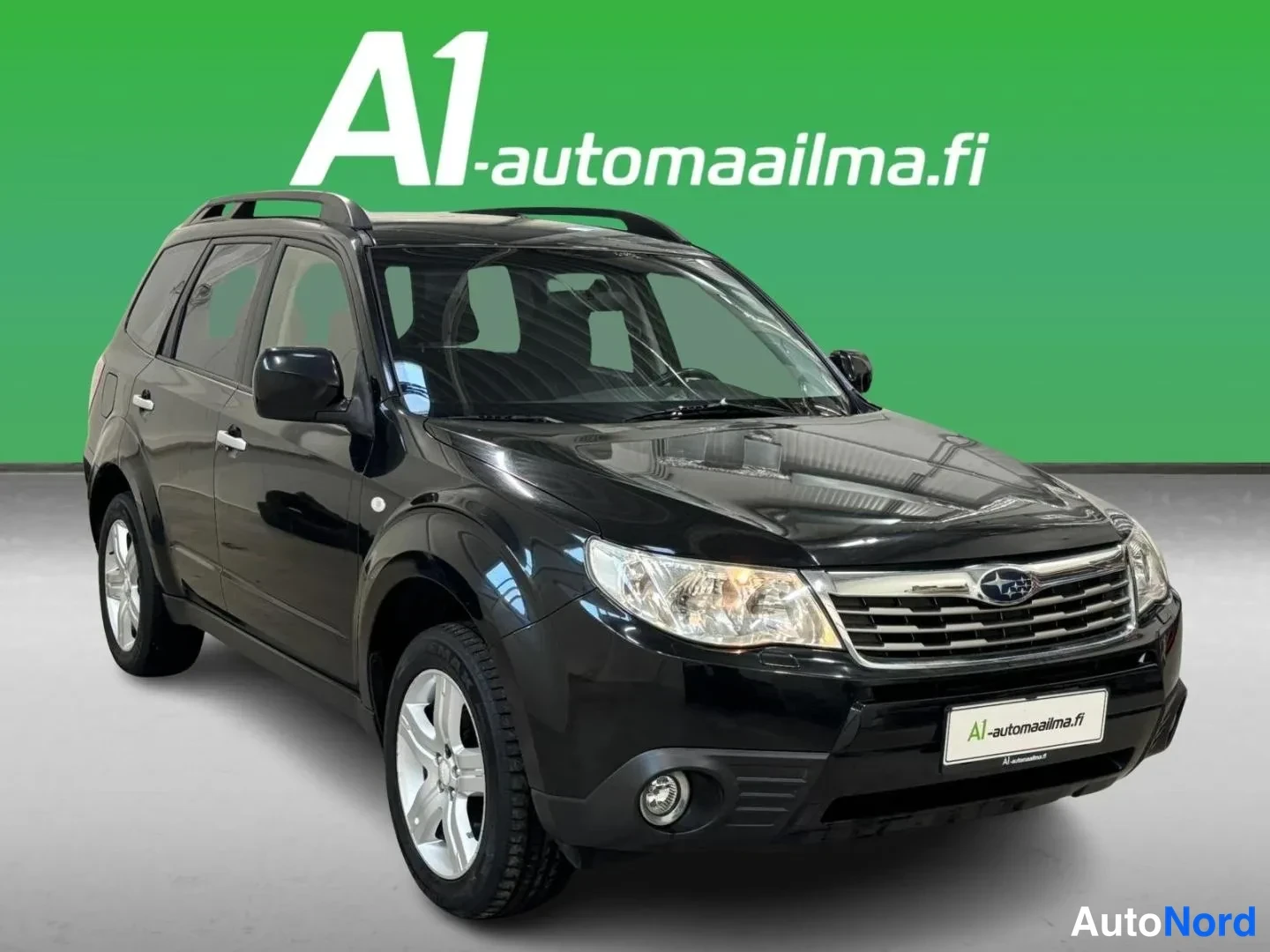 Etuviisto, nokka oikealle: Musta Subaru Forester kolmivartalokuvassa edestä oikealta, kattokaiteet ja hopeavanteet näkyvissä.