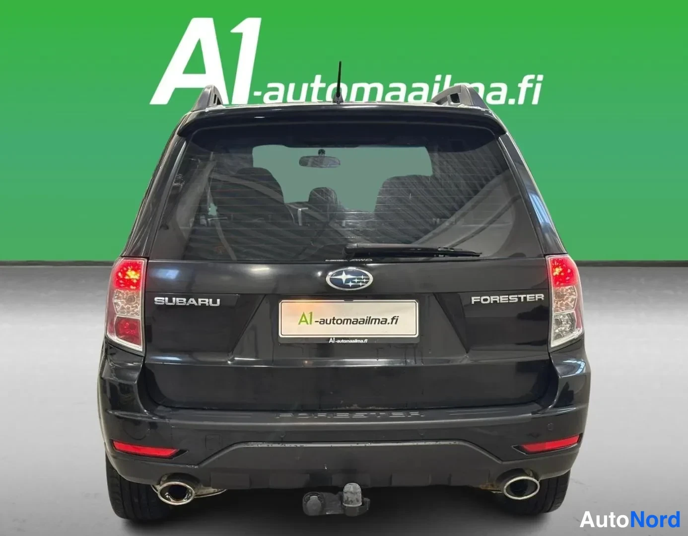 Takaa: Mustan Subaru Foresterin suora takanäkyymä: FORESTER-teksti, SUBARU-merkki, kattokaiteet, kaksinkertainen pakosarja ja perävaunukytkin näkyvät.