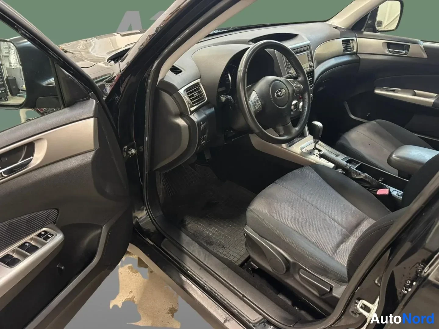 Edestä: Auton etuistuimilta kuvattu ohjaamo: kuljettajan ovii auki, Subarun ohjauspyörä, automaattivaihteiston valitsin ja keskiö console.