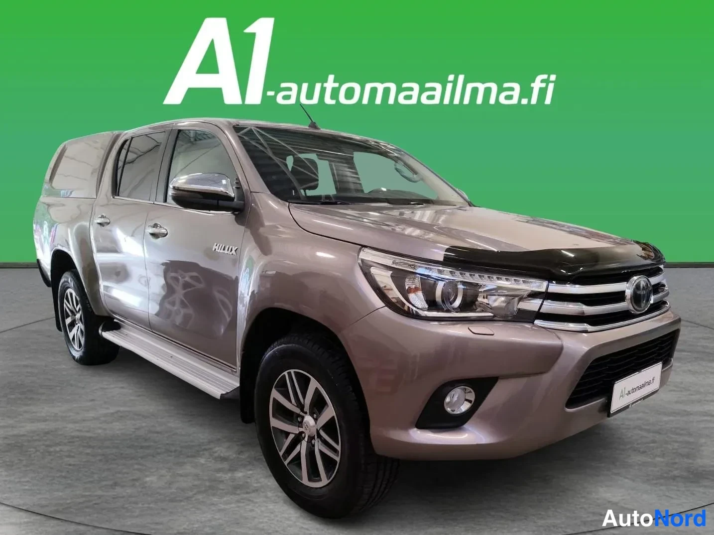 Etuviisto, nokka oikealle: Toyota Hilux Double Cab -noutoauton etukulmakuva, kevytmetallinvaalea runko, kromilamaanit ja tumma konepeltisuojus.