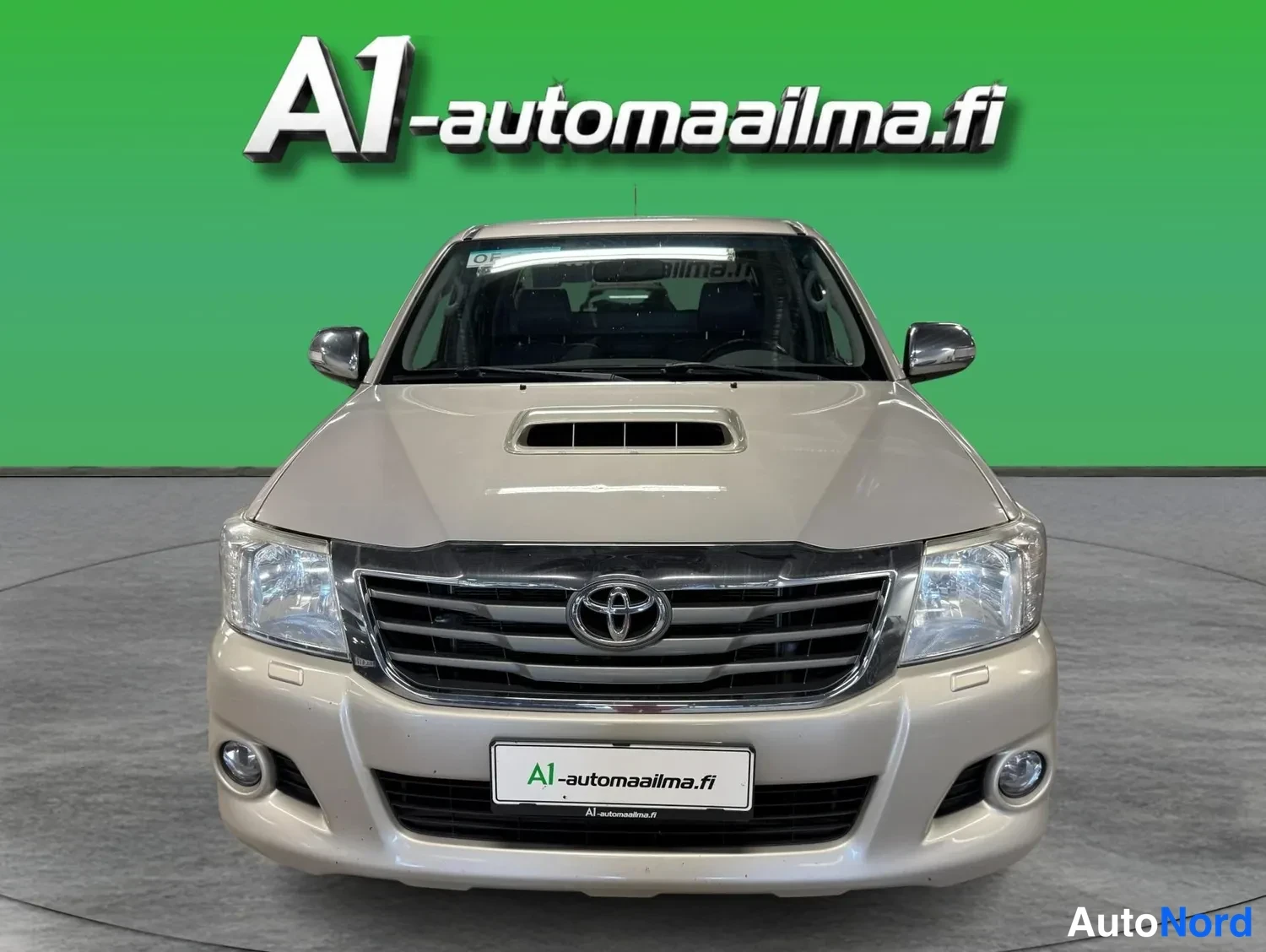 Edestä: Beige Toyota Hilux Double Cab -mallin keula edestä kuvattuna, sininen tausta.