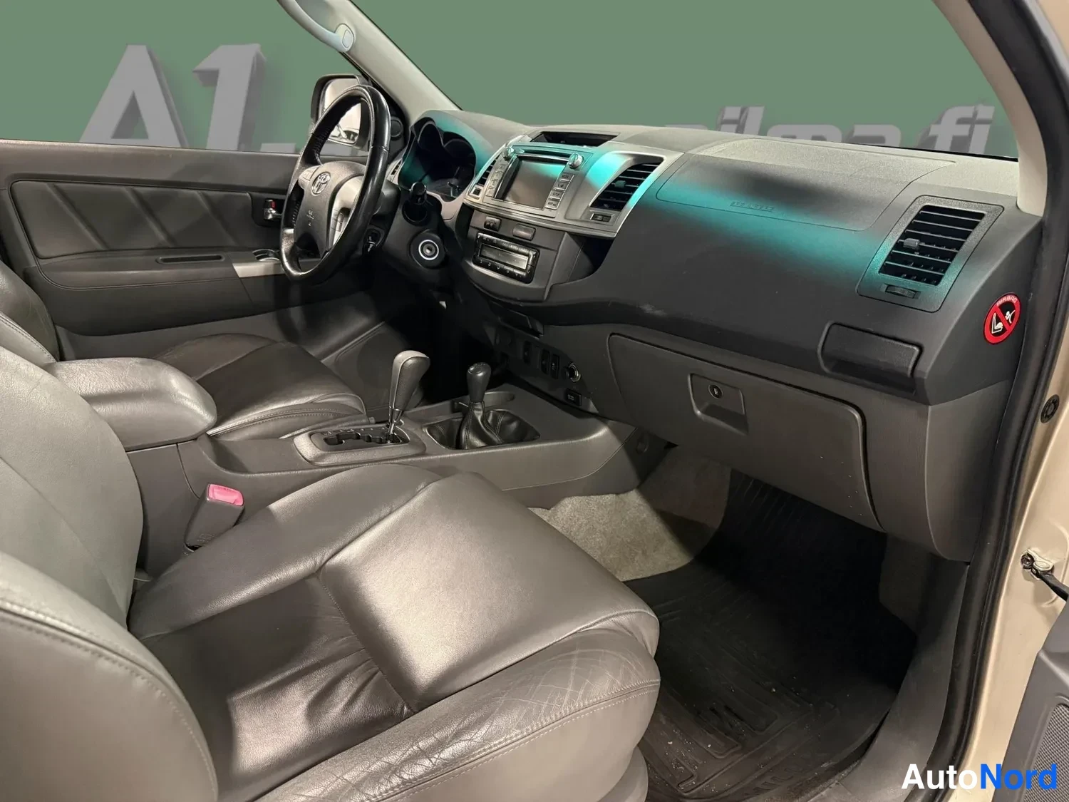 Etuviisto: Ohjaamosta matkustajan puolelta kuvattu näkymä: tummat nahkaistuimet, Toyota-merkkinen ohjauspyörä, radio-konsoli ja automaattivaihteen valitsin.