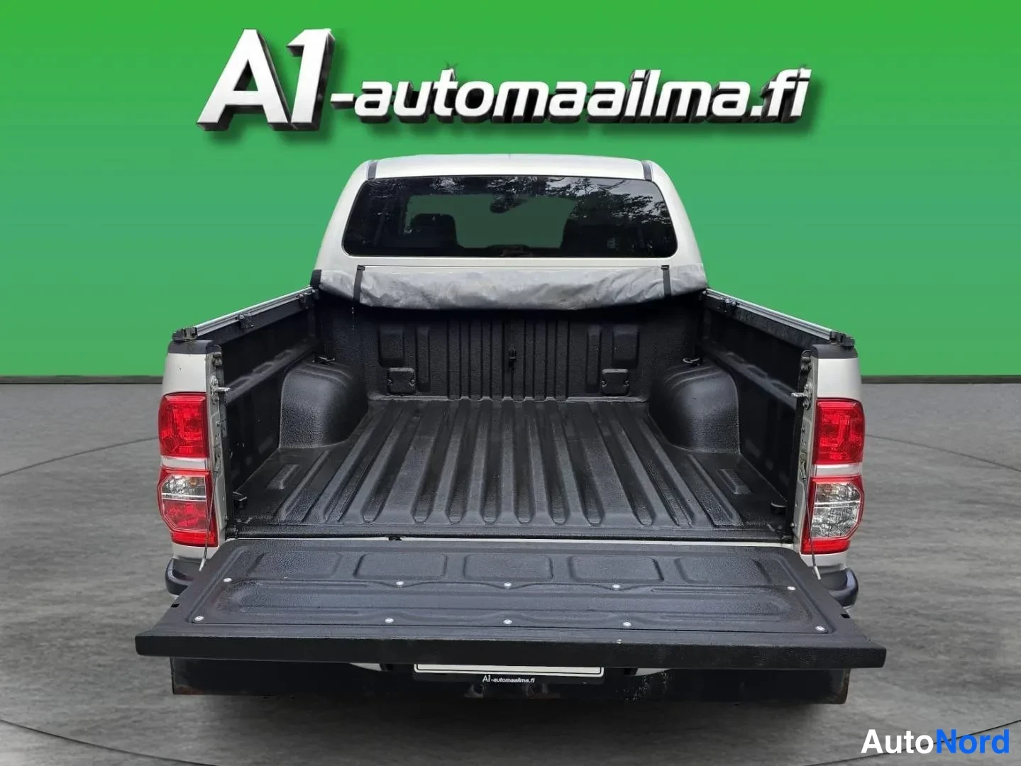 Takaa: Takaaparakuva lava-autosta: takaportti on avattu tasolle ja suoraan maahan, pehmeä pressu rullattuna kuutin etupuolelle; lavassa musta muovivuoraus.