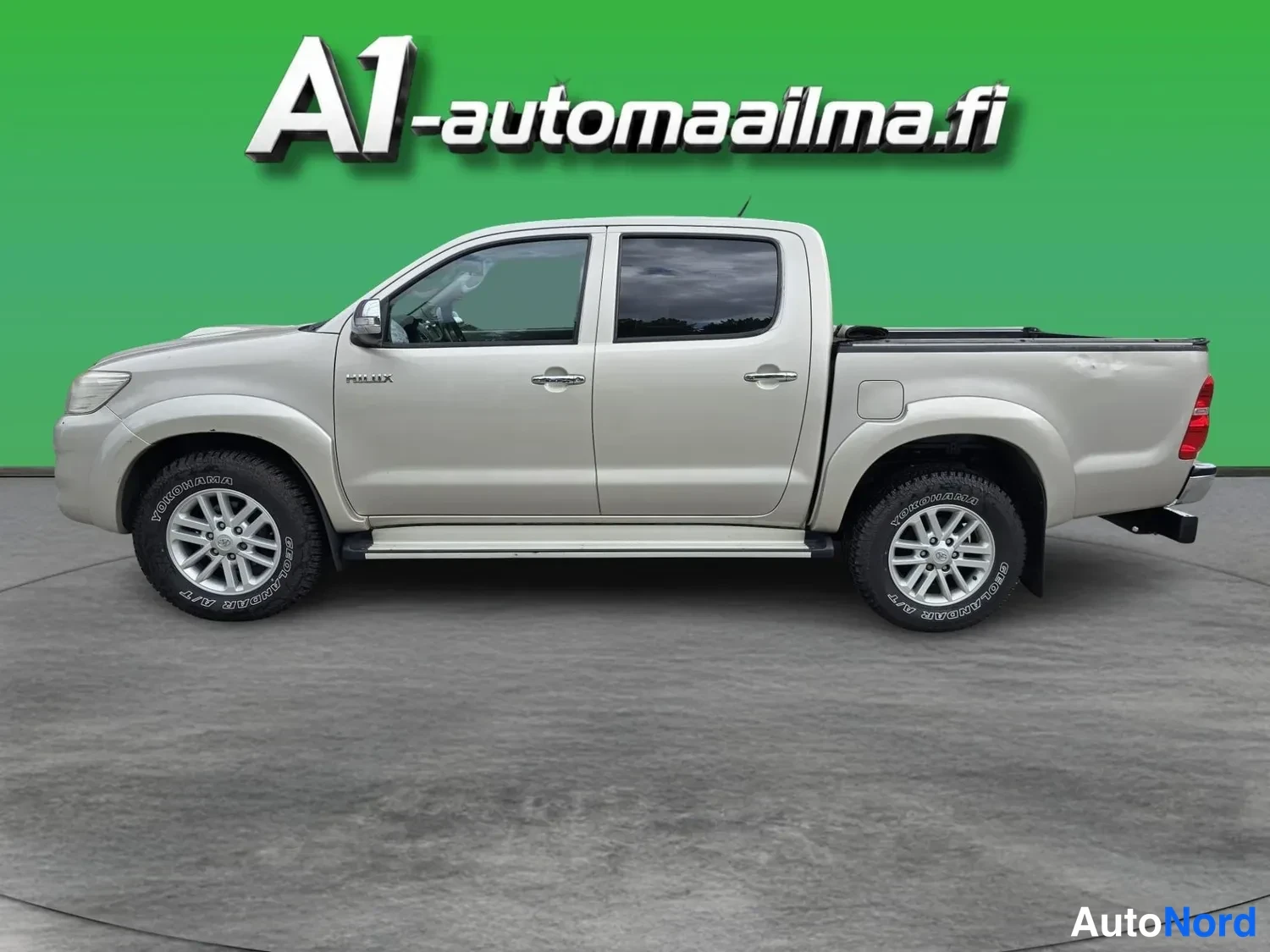 Takaviisto: Used pickup truck photographed as a rear three-quarter, nose pointing left, on grey floor with green backdrop. Body colour beige/gold metallic.