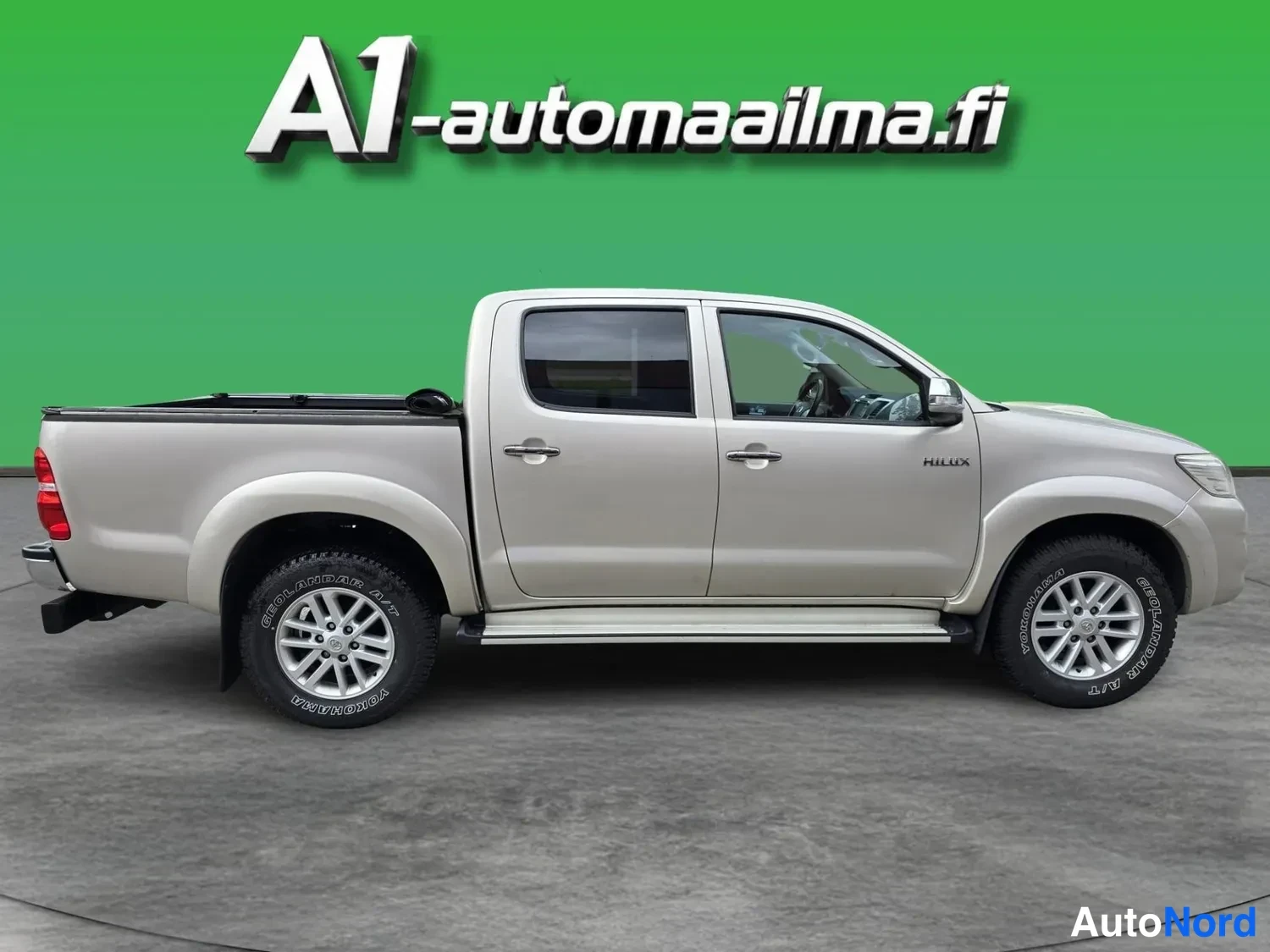 Etuviisto, nokka oikealle: Used pickup truck photographed as a front three-quarter, nose pointing right, on grey floor with green backdrop. Body colour beige/gold metallic.