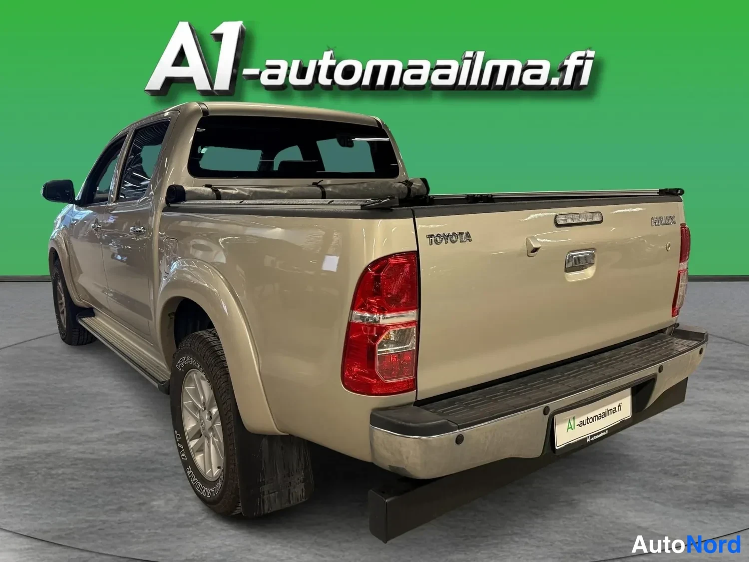 Takaviisto: Kultainen pickup takaa viistosti kuvattuna; lavan päällyskangas rullalla ja HOYOTA-HILUX-merkinnät peräportissa.