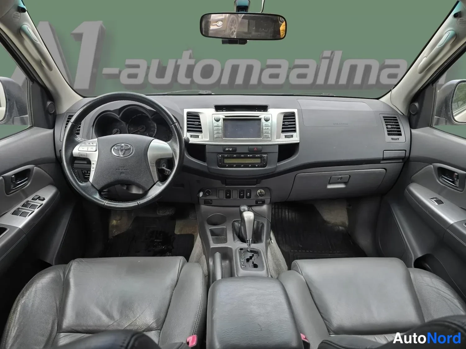 Etuviisto: Ohjaamosta suoraan eteenpäin kuvattu näkymä: tummanharmaat nahkaistuimet, Toyota-ohjauspyörä, kosketusnäyttö ja automaattivaihteen valitsin keskikonsolissa.