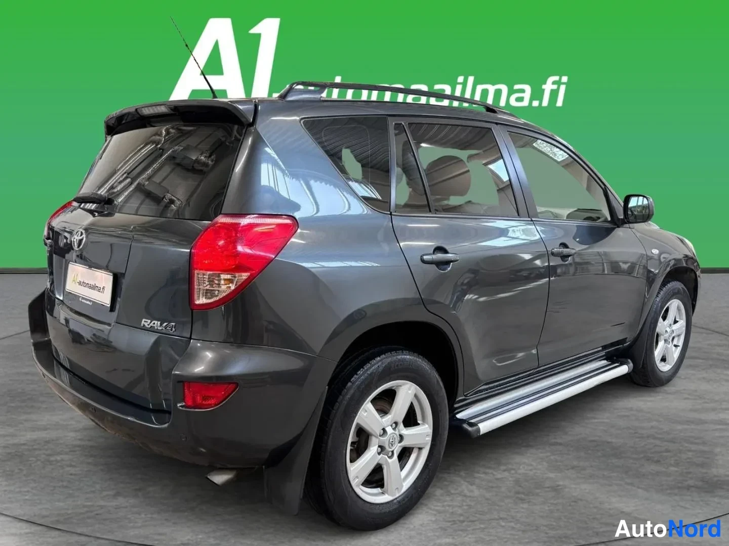 Takaviisto: Takanurkkikuva tummanharmaasta Toyota RAV4 -maasturista, kattokaiteet, takaviihdyttimen koroke ja sivulokiinateput näkyvät.
