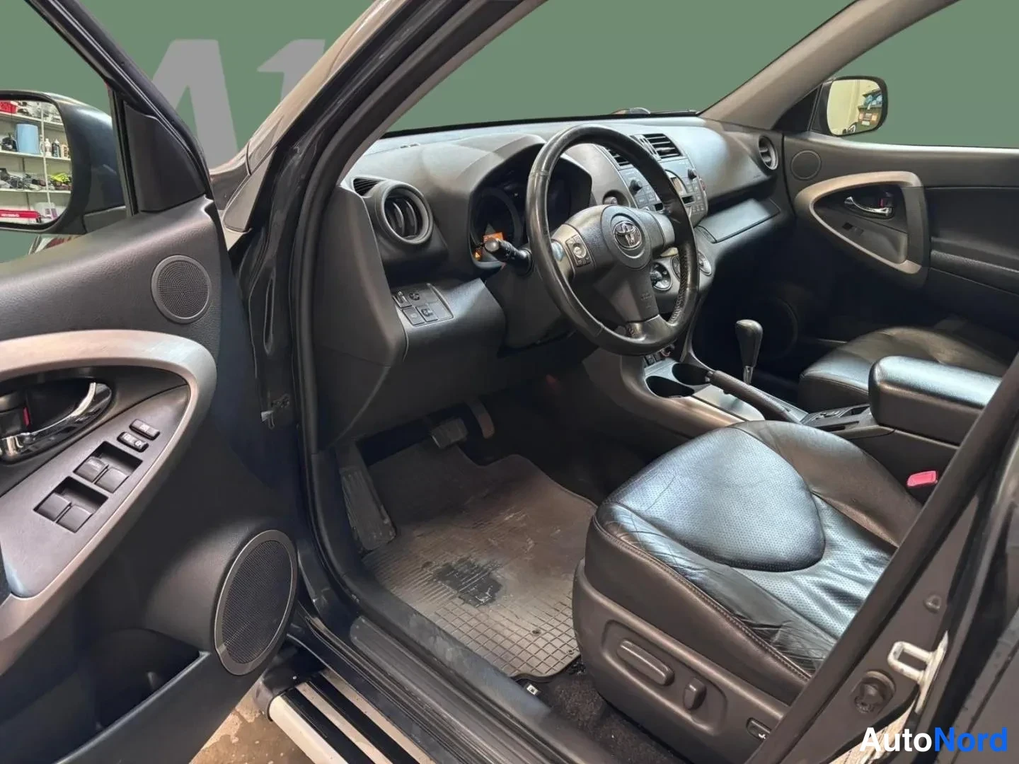 Muu: Avoimen kuljettajan oven näkymä tummaan ohjaamoon: Toyota-ohjauspyörä, vaihdin, nahkaverhoillut etuistuimet ja oven ikkunakytkimet.