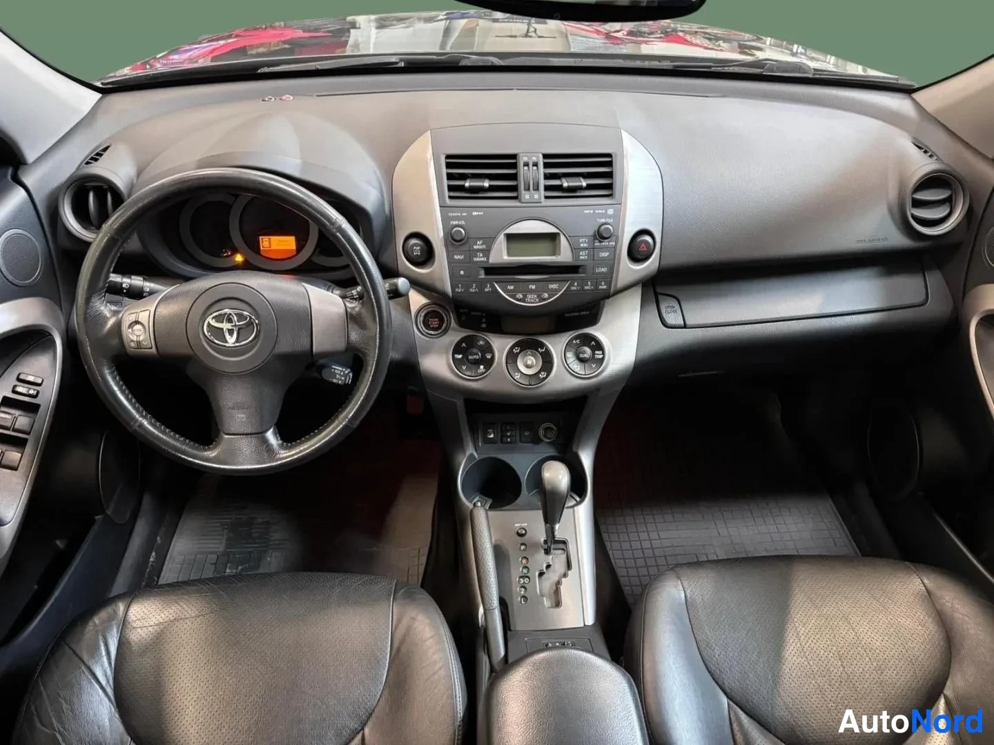 Muu: Toyota RAV4:in ohjaamo nähtynä kojelaudan yli: tumma mittaristo ja Toyota-ohjauspyörä, keskellä radio ja automaattivaihteen valitsin, mustat nahkaistuimet etual