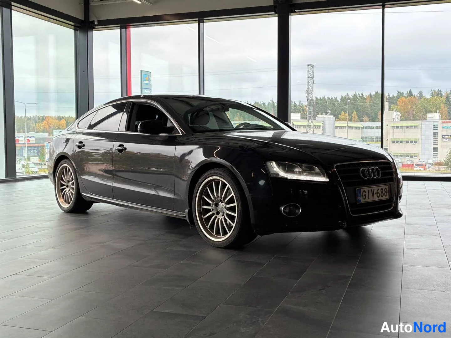 Etuviisto, nokka oikealle: Mustan Audi Sportbackin etukulma nähtynä oikealta edestä sisätiloissa.