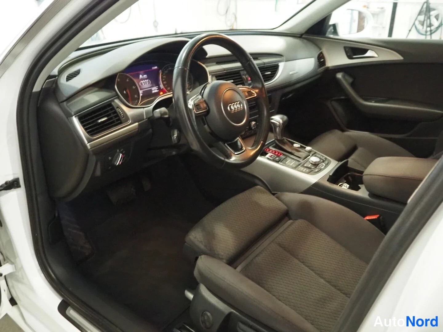 Muu: Audi A6:n sisätilat: ohjauspyörä ja kojelauta vasemmalla, automaattivaihteen valitsin ja keskikäyttölaitteet, tummat kangasistuimet.