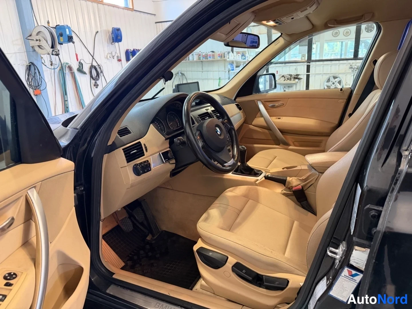 Muu: Kuva BMW X3:n sisätilasta kuljettajan ovesta: beigeä nahkaa penkeissä, tumma kojelauta, BMW-ohjauspyörä, manuaalivaihteen keksi ja koroke, tallin työtilat näkyv