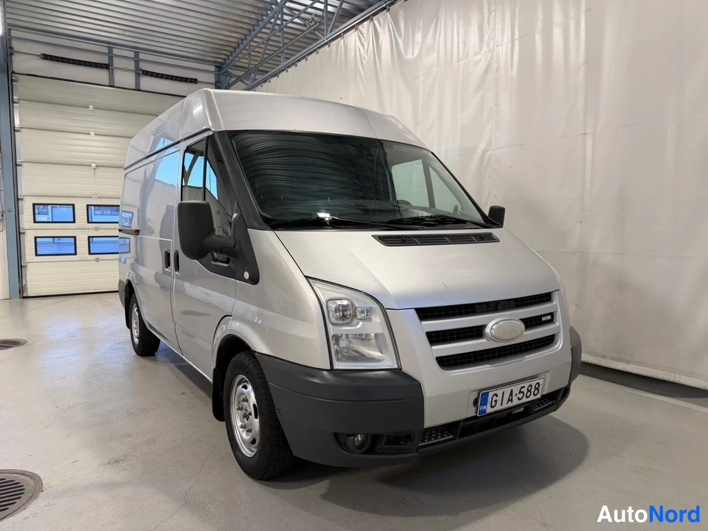 Etuviisto, nokka oikealle: Hopeanvärinen Ford Transit -pakettiauto kuvattu kolmanneskulmasta keula oikealla hallissa; rekisterikilpi etupuskurissa.
