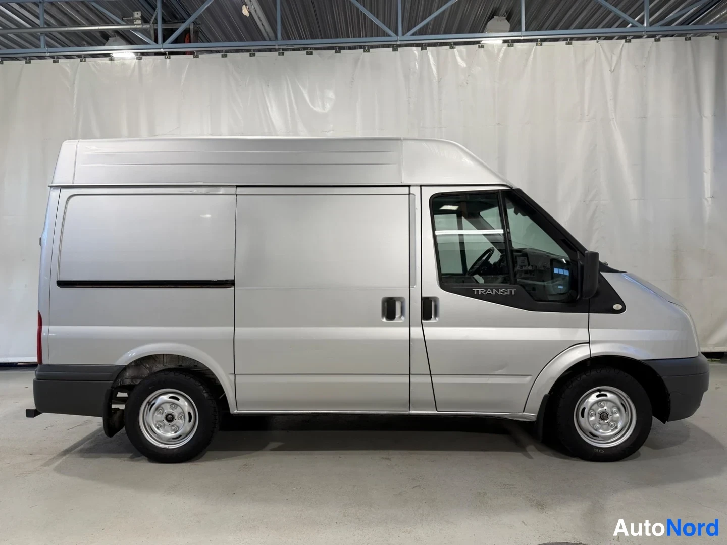 Sivulta: Hopeanvärinen Ford Transit -pakettiauto sivusta oikealta kuvattuna hallissa; TEXT-näkymä liukuvista takaovista ja korkeasta katosta.
