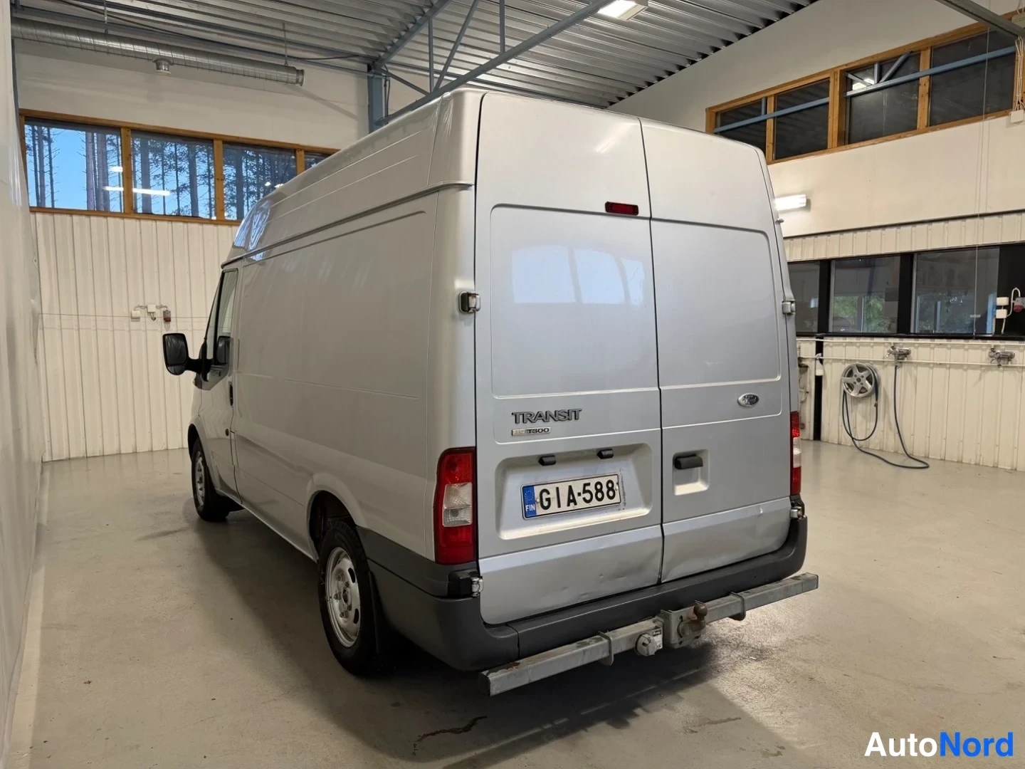 Takaviisto: Hopeanvärinen Ford Transit -pakettiauto takaviisto oikealta hallissa; rekisterikilpi ja takaovekyljet luvut 'TRANSIT' ja '350'.