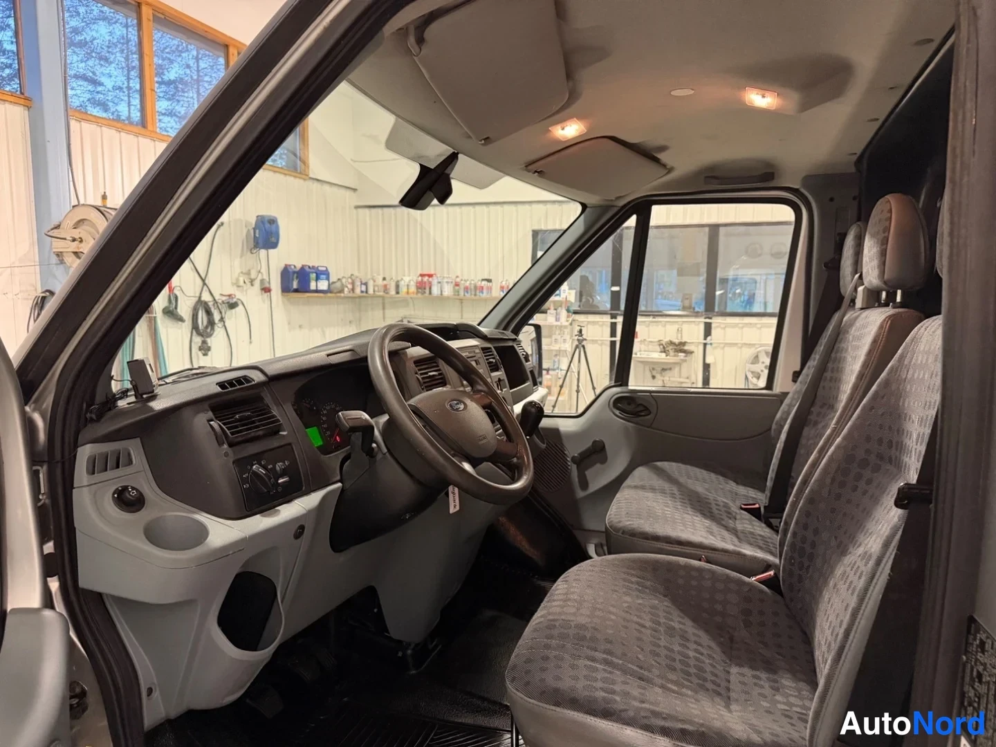 Muu: Ford Transitin ohjaamo sisältä: ohjauspyörä, tumma kojelauta ja harmaankuvioiset istuimet.