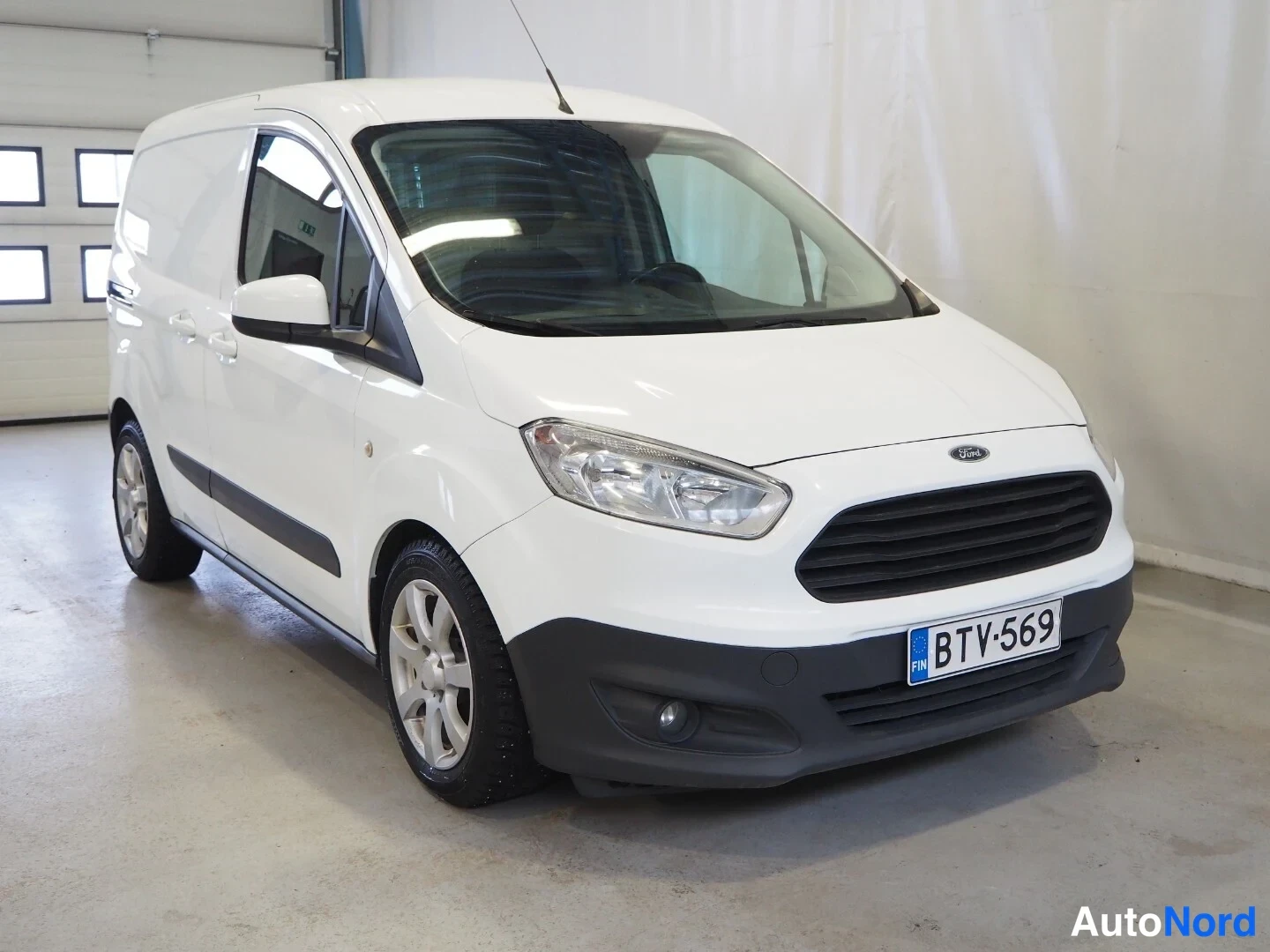 Etuviisto, nokka oikealle: Valkoinen Ford Transit Courier -pakettiauto etuosa vasemmalta kolmannelta kuvakulmalla, rekisteritunnus.