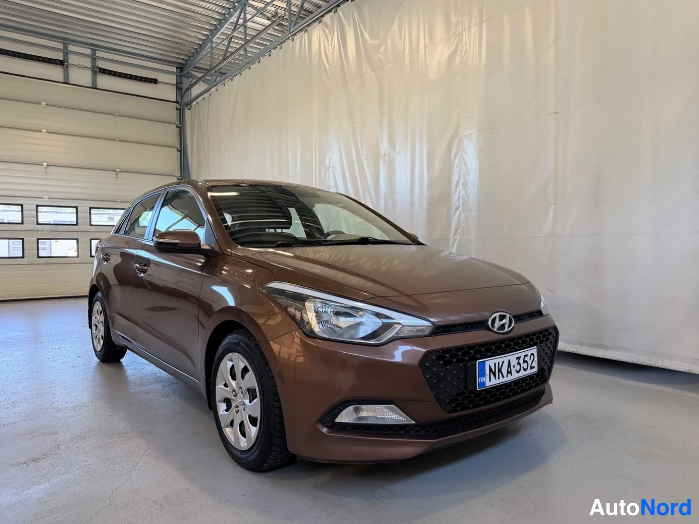 Etuviisto, nokka oikealle: Ruskea Hyundai i20 -henkilöauto etuosa vasemmalle kääntyneenä kuvattuna sisätiloissa, rekisteritunnus.