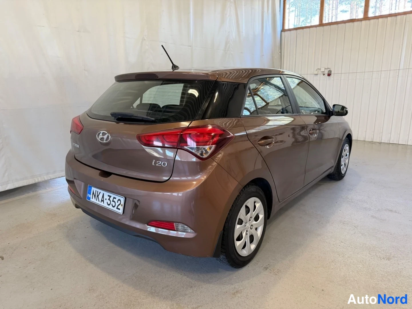 Takaviisto: Ruskea Hyundai i20 -henkilöauto takaa oikealta sivulta kuvattuna sisätiloissa, takavalot ja rekisteritunnus näkyvissä.