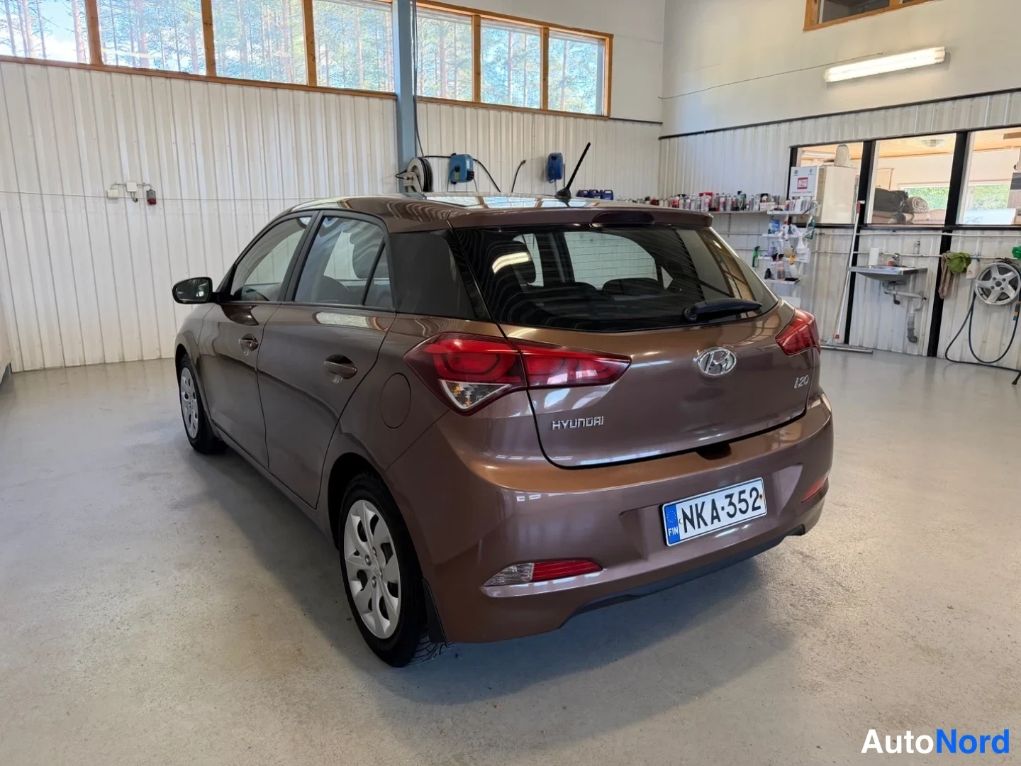 Takaviisto: Ruskea Hyundai i20 -henkilöauto takaa vasemmalta sivulta kuvattuna huoltotilassa, takaoven ja rekisteritunnuksen näkyessä.