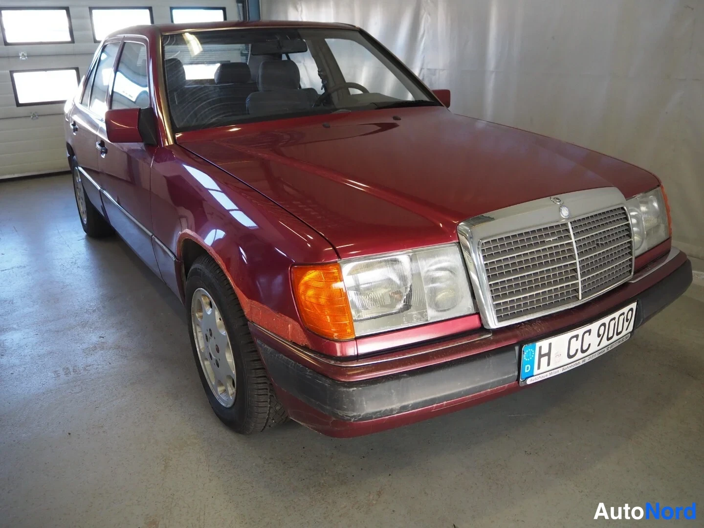 Etuviisto, nokka oikealle: Punaviininpunainen Mercedes-Benz W124 -sedan etu- ja sivukulmasta hallissa, etusäleikkö ja ajovalot näkyvät, rekisterikilpi H CC 9009.