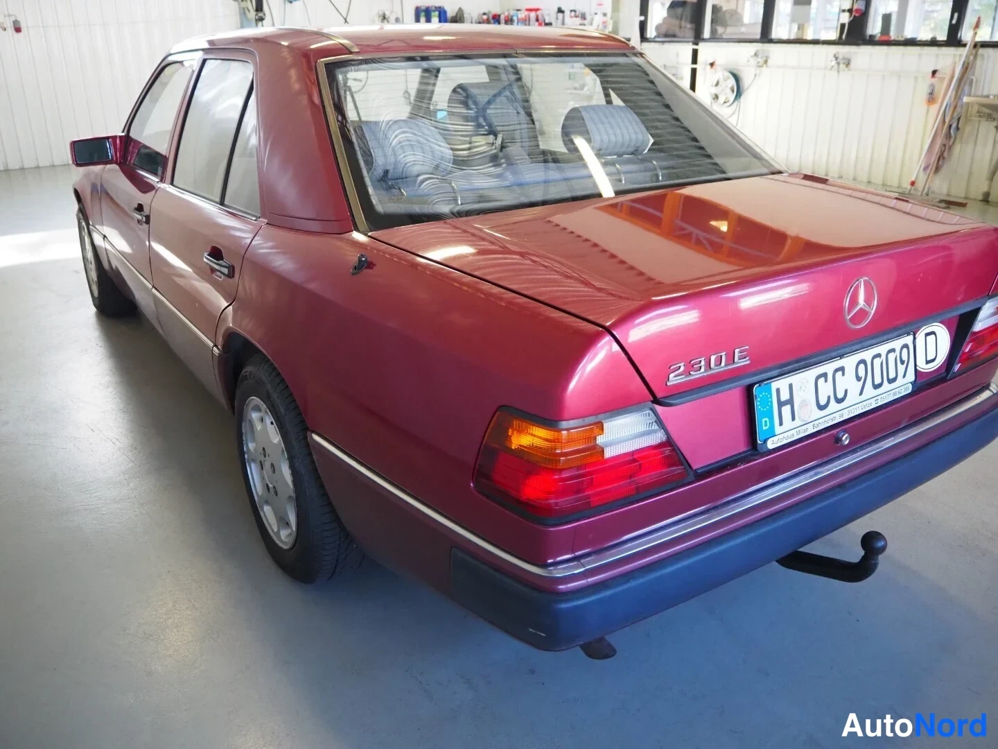 Takaviisto: Punaviininpunainen Mercedes-Benz W124 -sedan taka- ja sivukulmasta autotallissa, takavalo ja peräosat näkyvissä, takana rekisterikilpi H CC 9009.