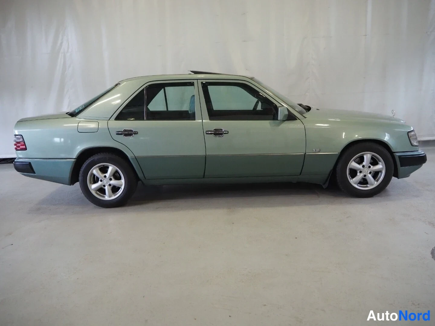 Sivulta: Mercedes-Benz W124-sedanin sivuprofiili sisätiloissa, vihreä kori, neljä ovikäsislintaa ja alumiinivanteet näkyvissä.
