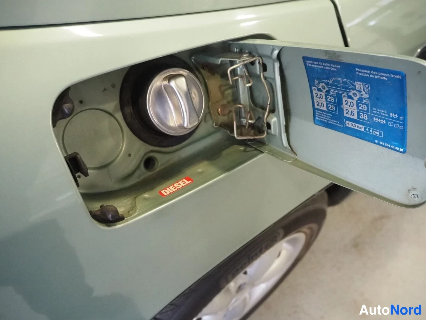 Sivulta: Vaaleanvihreä auton avattu tankin luukku metallisella tankin kannella, Diesel-tarra ja täydennyspainetaulukko.