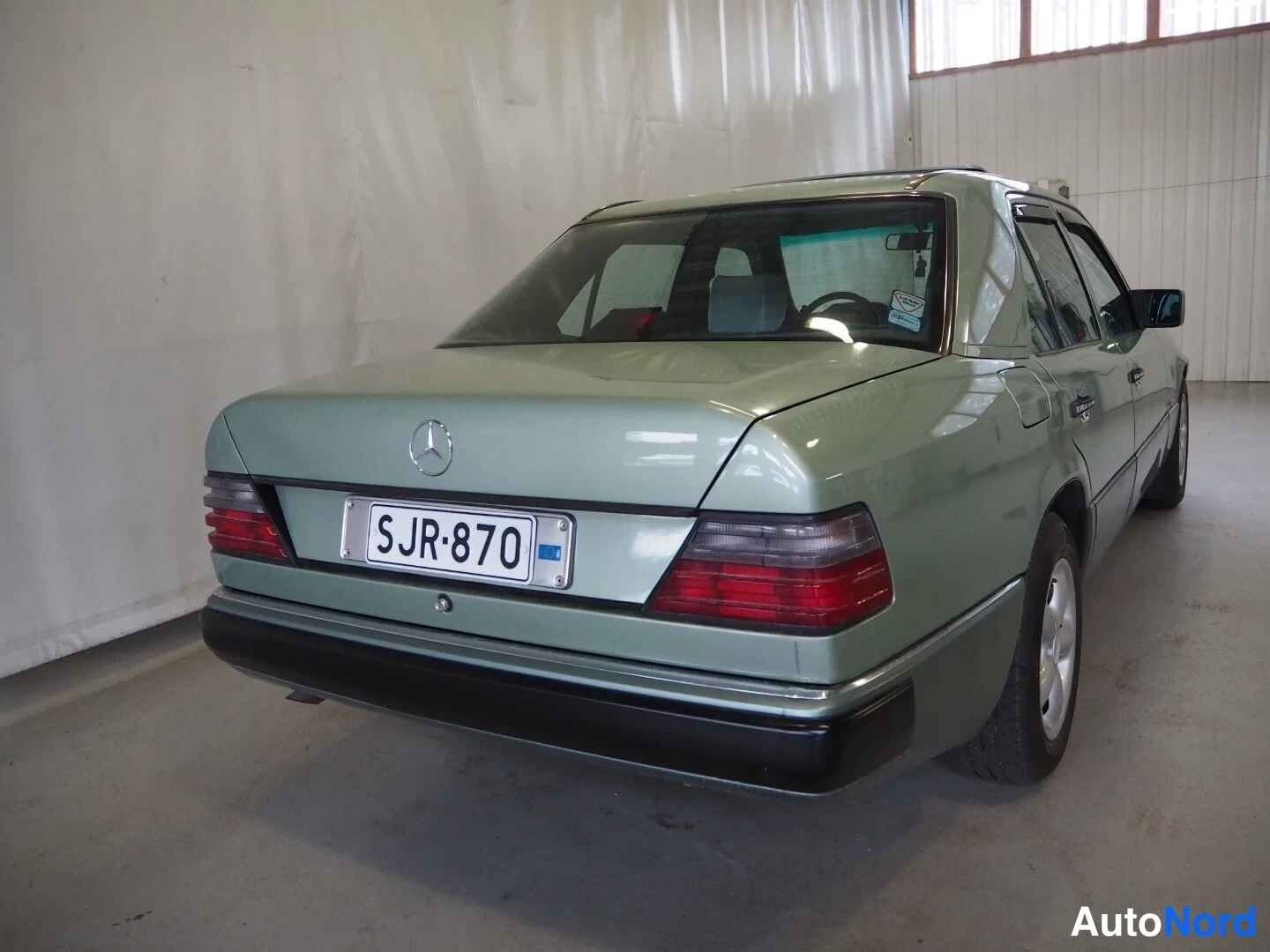 Takaviisto: Mercedes-Benz W124-sedanin takakulma sisätiloissa, vihreä kori, rekisterikilpi ja Mercedes-tähti kannessa.