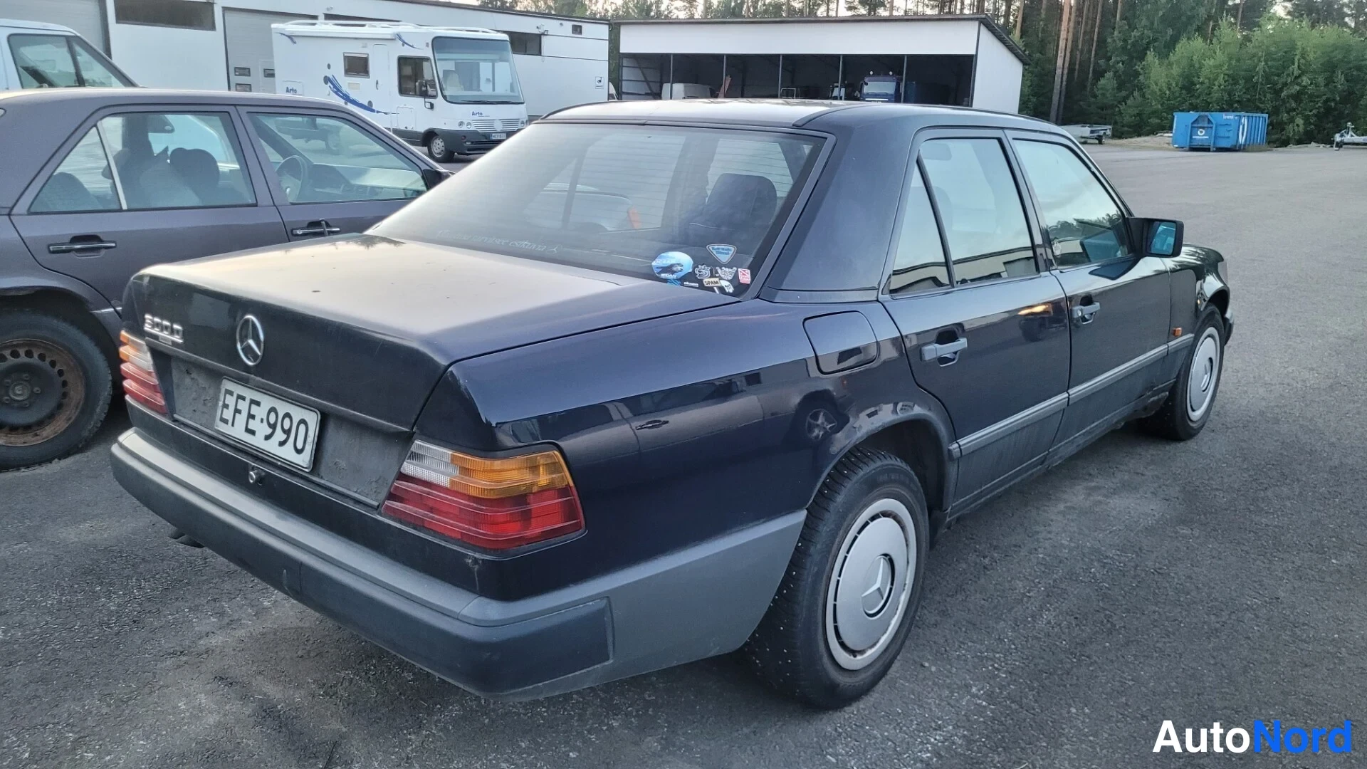 Takaviisto: Tummansininen Mercedes-Benz-sedan takapäältä vasemmasta neljäsosasta, rekisteritunnus, takalokasuojassa peili ja takaluukun tarrat.
