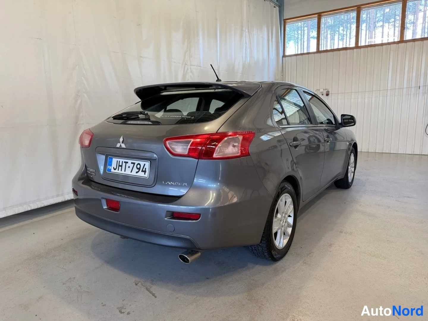 Takaviisto: Tummanharmaan Mitsubishi Lancer -auton takakulma, takapuoli ja oikea sivu näkyvät, rekisterikilpi.