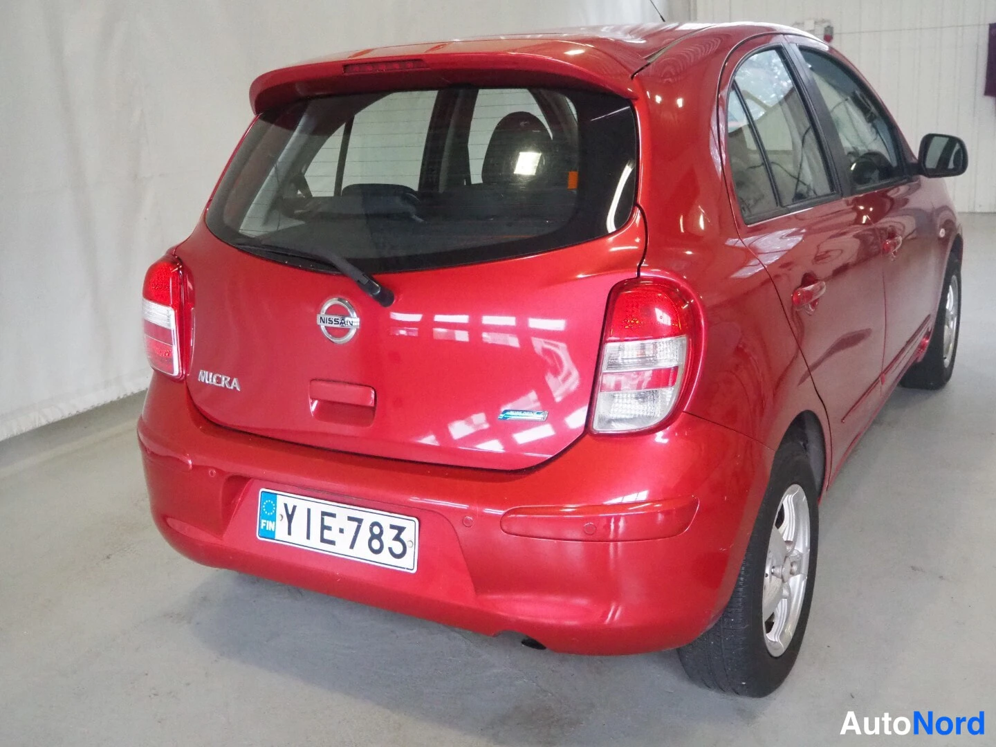 Takaviisto: Punaisen Nissan Micran takakulma, MICRA-tunnus, Nissan-merkki ja rekisterikilpi näkyvät sisätilassa.