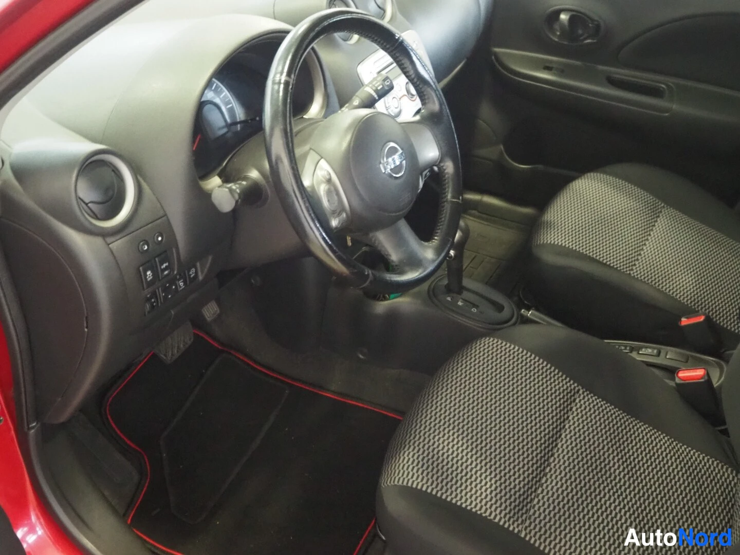Edestä: Auton etusissa: Nissan-ohjauspyörä, tumma kojelauta, manuaalivaihteisto ja punaista reunustaa pitävät lattiamatot.