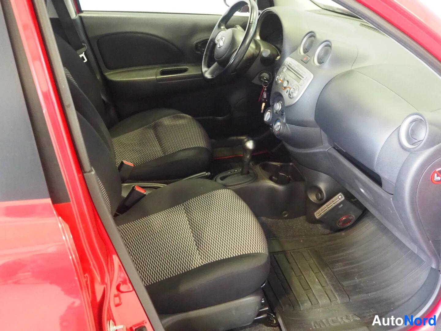 Edestä: Kuljettajan puolen sisätila: Nissan-ohjauspyörä, tummat kankaat istuimet ja lattiamatto, johon on painettu sana MICRA.