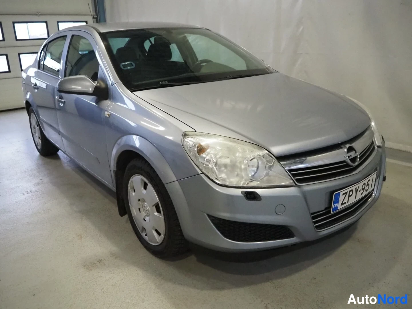 Etuviisto, nokka oikealle: Hopeanharmaa Opel Astra -sedan näkyvä etusivu ja oikea kylki edestä oikealta kuvattuna, rekisteritunnus.
