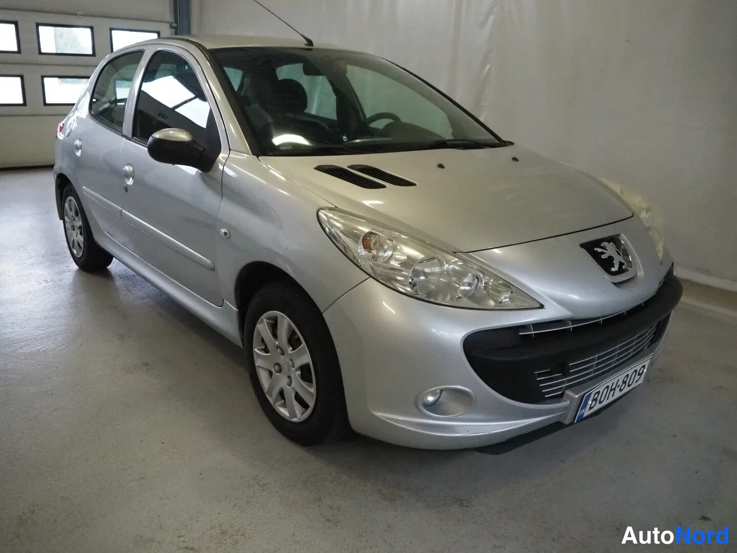 Etuviisto, nokka oikealle: Hopeanvärinen viisiovinen Peugeot 206+ -henkilöauto kuvattu kolmasosakulmasta edestä oikealta sisätilassa.