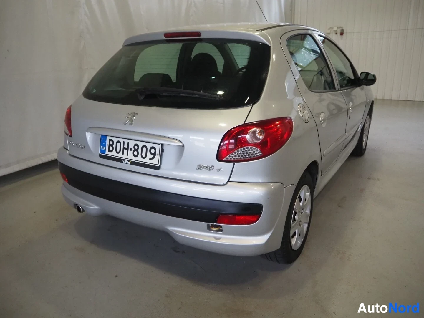 Takaviisto: Hopeanvärinen Peugeot 206+ -henkilöauto kolmasosakulmasta takaa vasemmalta, rekisterikilpi.