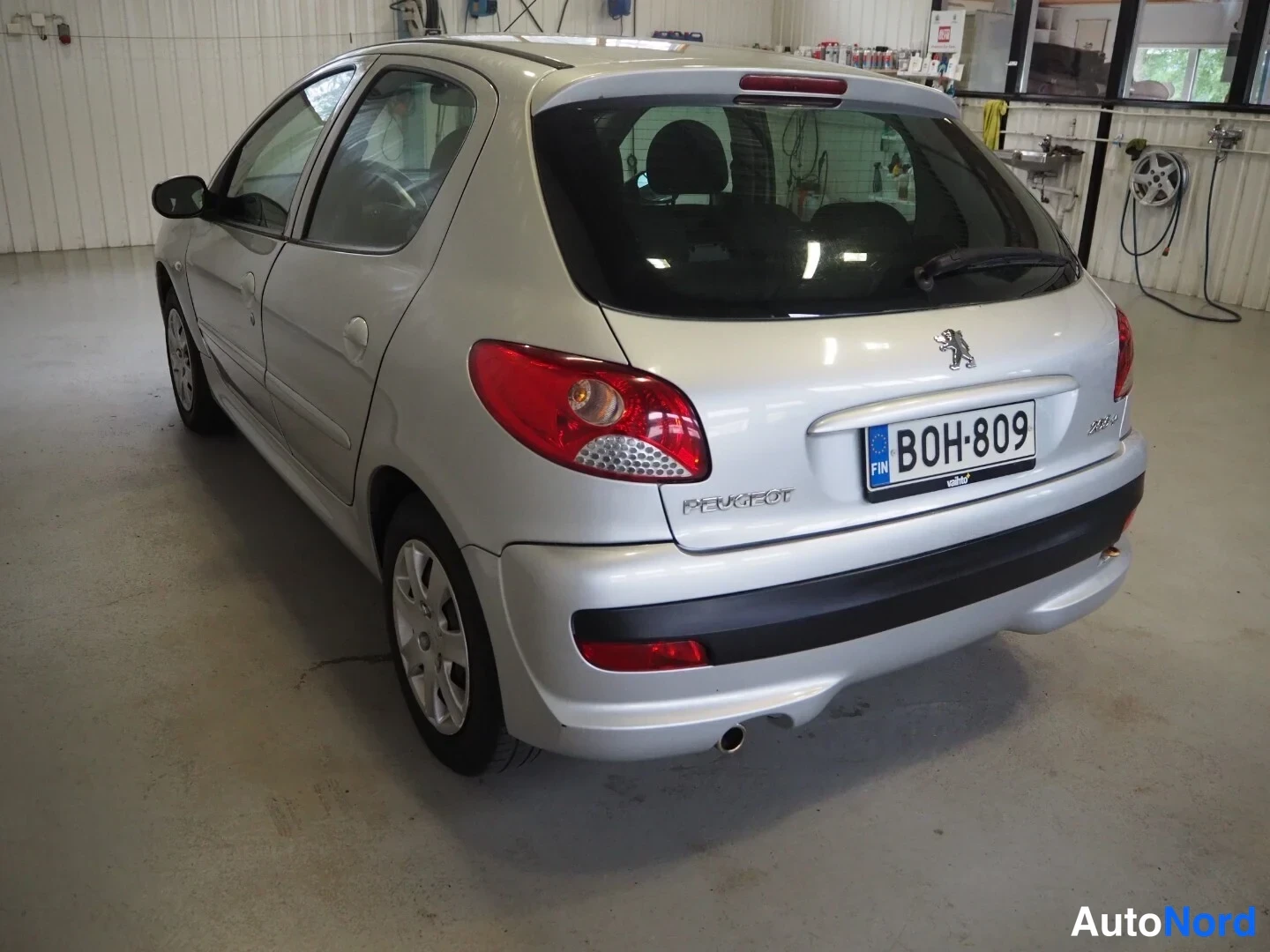 Takaviisto: Hopeanvärinen Peugeot 206+ -henkilöauto takaa vasemmalta korjaamotilassa, rekisterikilpi.