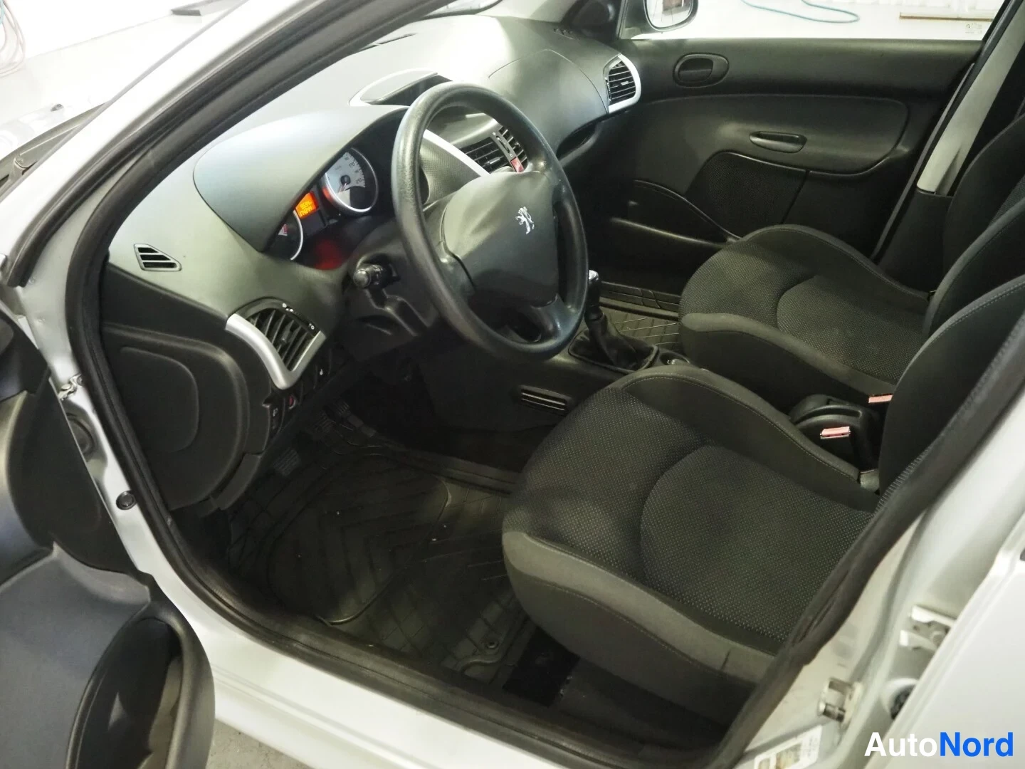 Muu: Peugeot 206:n matkustamon kuljettajan puoli: ohjauspyörä Peugeot-logolla, tummat kankaat etumatkustajan istuin, penkki, mattosarja ja manuaalivaihteiston vaihde