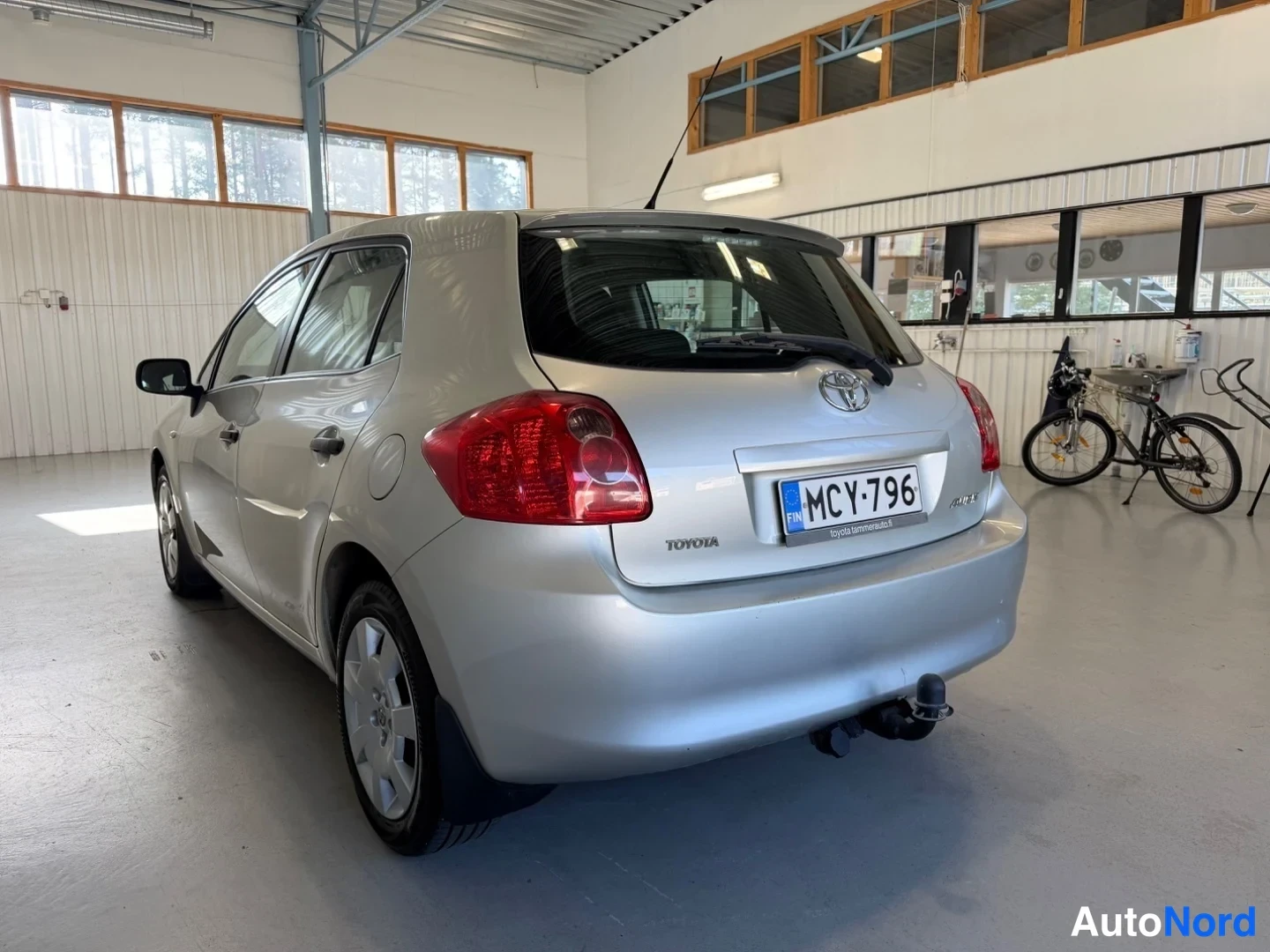 Takaviisto: Hopeanvärinen Toyota Auris takaa kolmannesvälikulmasta vasemmalle, rekisterikilpi, nosturitilan takaseinä näkyy, hallitila.