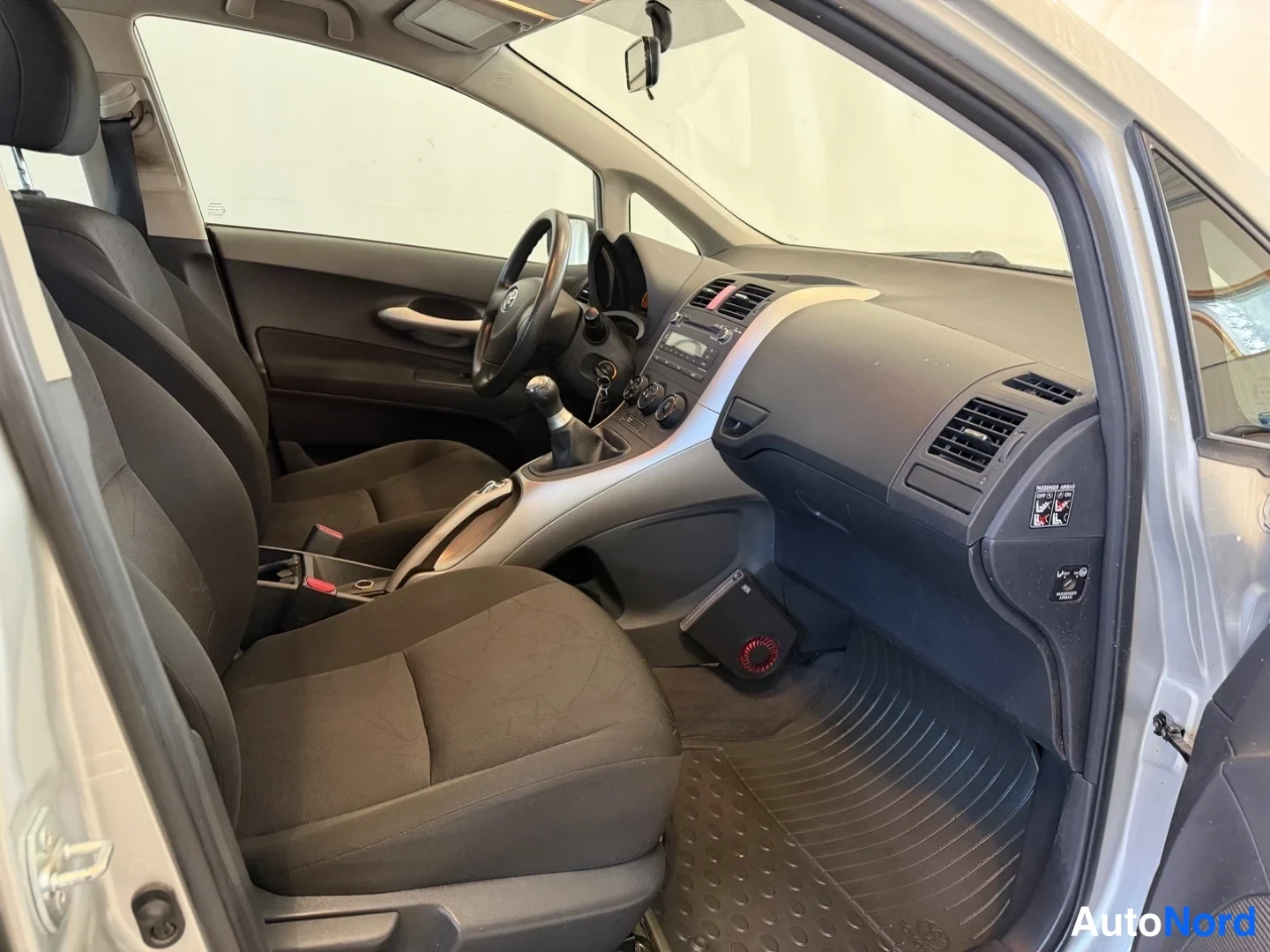 Muu: Toyota Aurisin etuistuimet ja kojelauta katsottuna matkustajan oven aukosta; kangasistuimet, manuaalivaihteovipu ja radiatorimatto näkyvät.