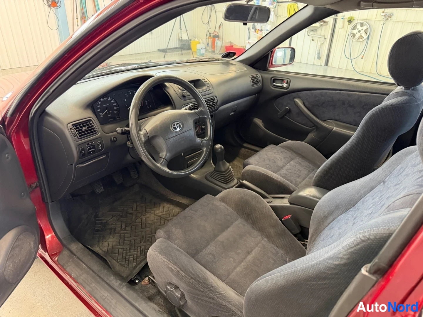 Ohjaamo: Driver side interior of the car showing dashboard, steering wheel with Toyota logo, gear lever, door panel, and front seats.